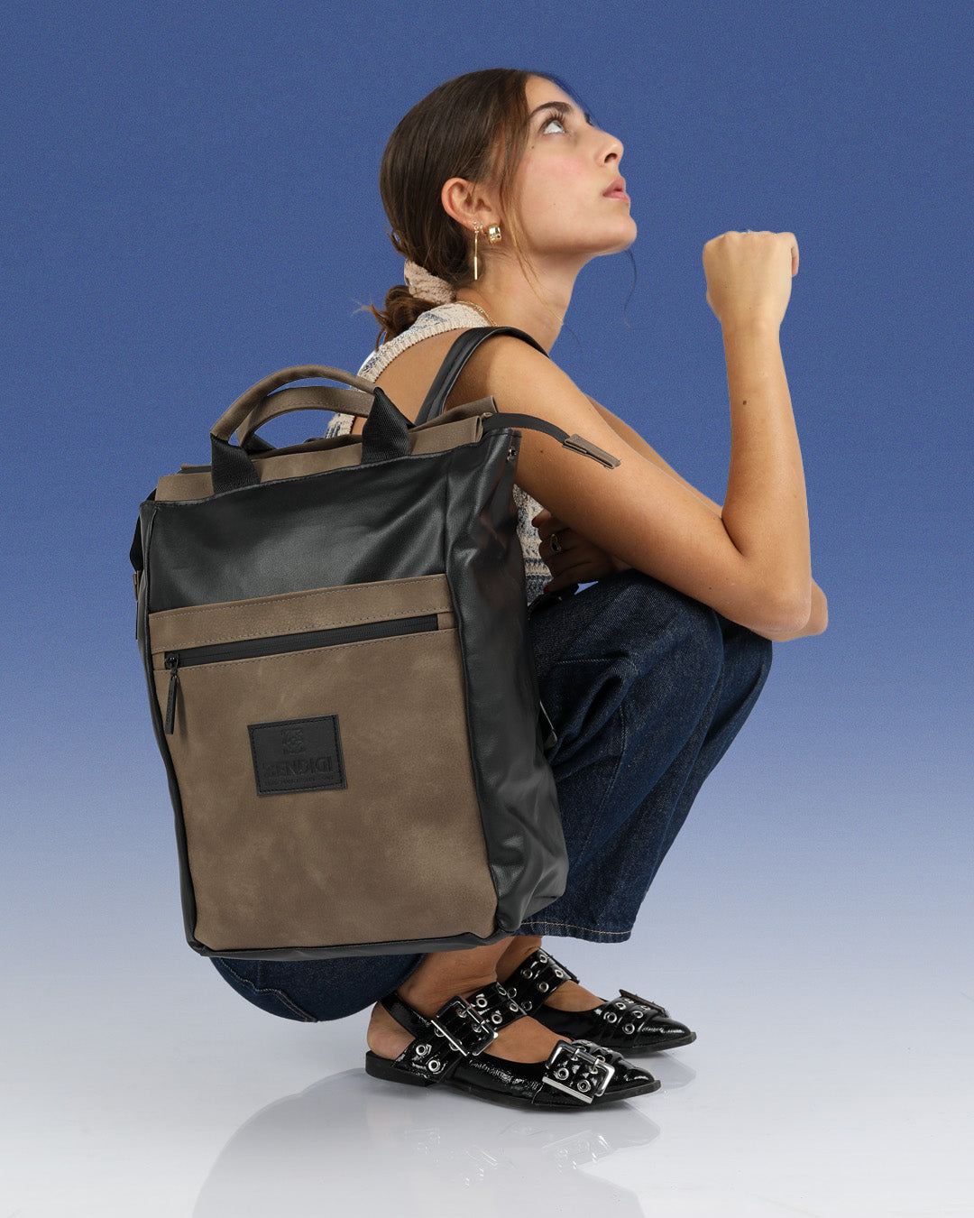 דוגמנית יושבת ועל הכתף שלה  תיק מיני מילאנו שחור מבריק וקפה, תיק עוצר נשימה ביופיו, תיק גב טבעוני למחשב נייד עד 14״, תיק גב לאוהבות הסטייל והיופי, תיק איכותי שמחזיק מעמד שנים קדימה.