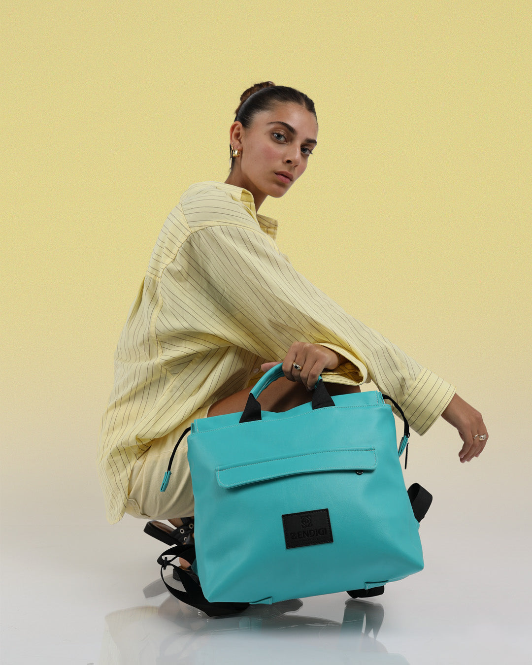 תיק גב מדריד טורקיז מיקונוס, מוחזק ביד על דוגמנית, ניתן לראות את גודלו באופן פורפורציונלי לדוגמנית, תיק גב טורקיז למחשב, תיק לנשיאה בידיים.