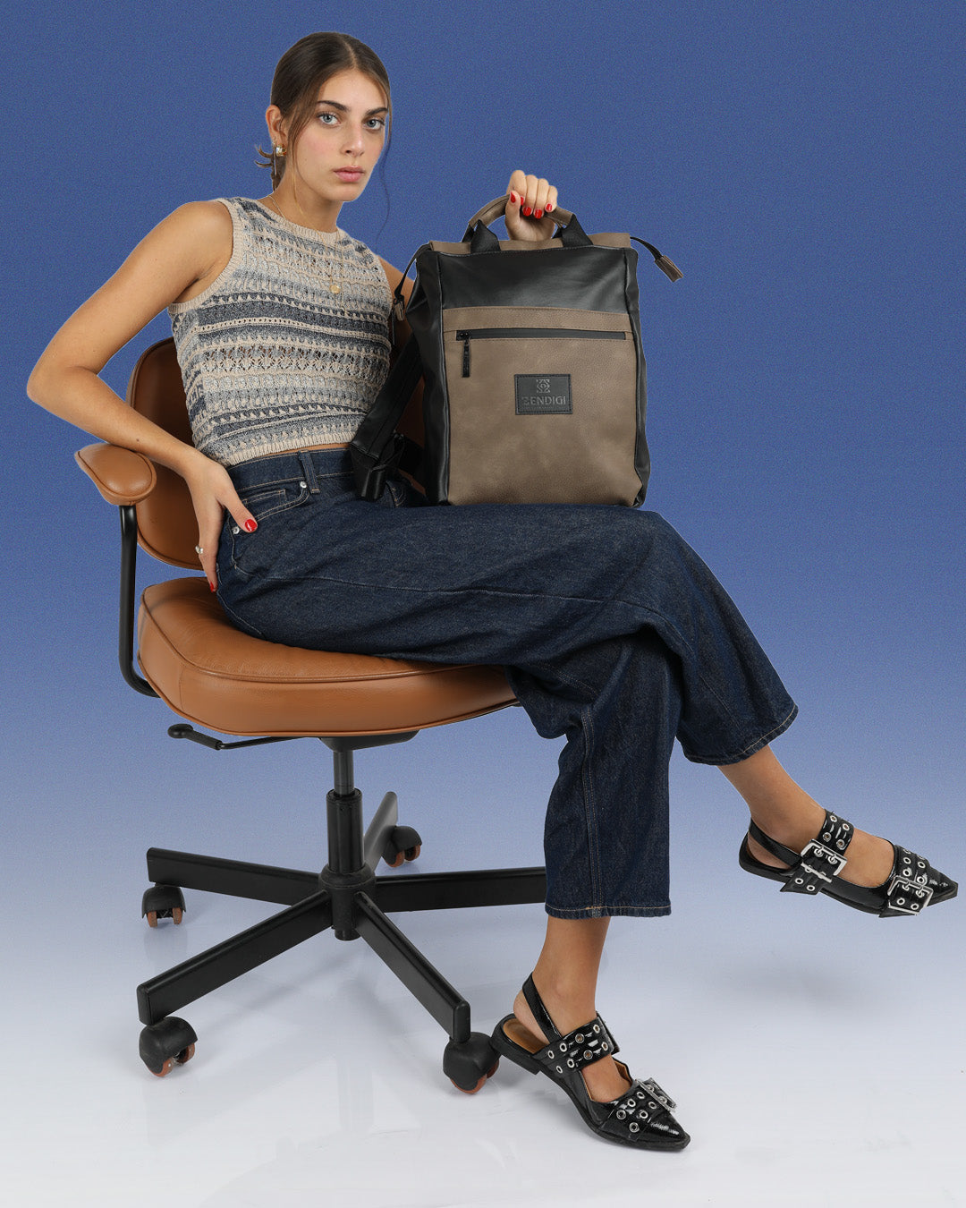דוגמנית יושבת על כיסא לימודים מחזיקה בידית הנשיאה את התיק מיני מילאנו שחור מבריק, תיק למחשב 14״ עם תא מרופד בעיצוב נקי וקלאסי עם נגיעות אורבניות, תיק לסטודנטים, תיק לעבודה, תיק ליום מחוץ לבית