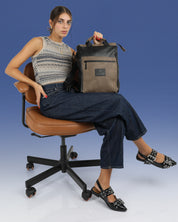 דוגמנית יושבת על כיסא לימודים מחזיקה בידית הנשיאה את התיק מיני מילאנו שחור מבריק, תיק למחשב 14״ עם תא מרופד בעיצוב נקי וקלאסי עם נגיעות אורבניות, תיק לסטודנטים, תיק לעבודה, תיק ליום מחוץ לבית