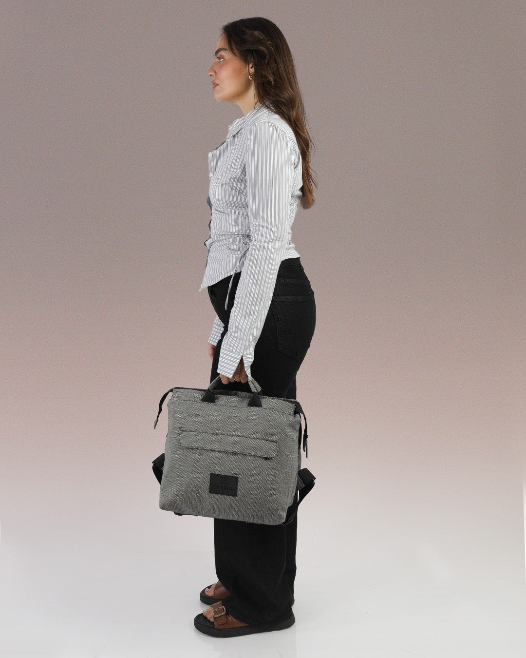 תיק גב מדריד, מוחזק ביד על דוגמנית, ניתן לראות את גודלו באופן פרופורציונלי לדוגמנית, תיק גב אפור למחשב, תיק לנשיאה בידיים.